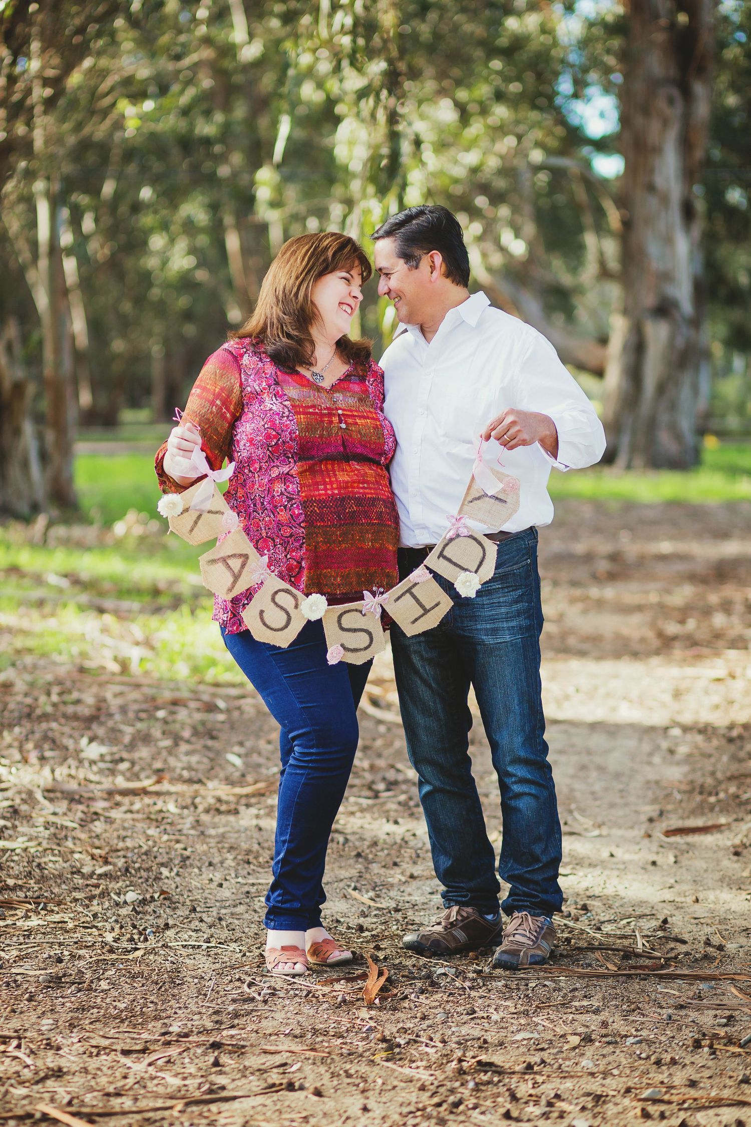 Couple poses with 'IT'S A GIRL' banner during forest maternity photoshoot.