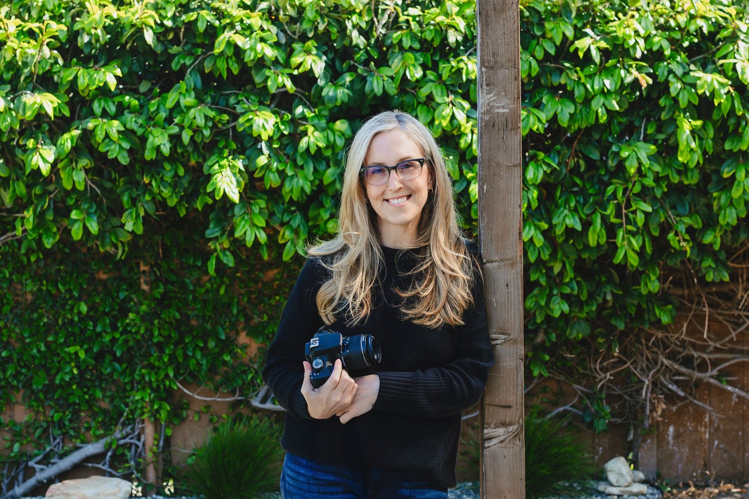 A person in a black sweater holding a small black dog stands in front of a lush green vine-covered wall.