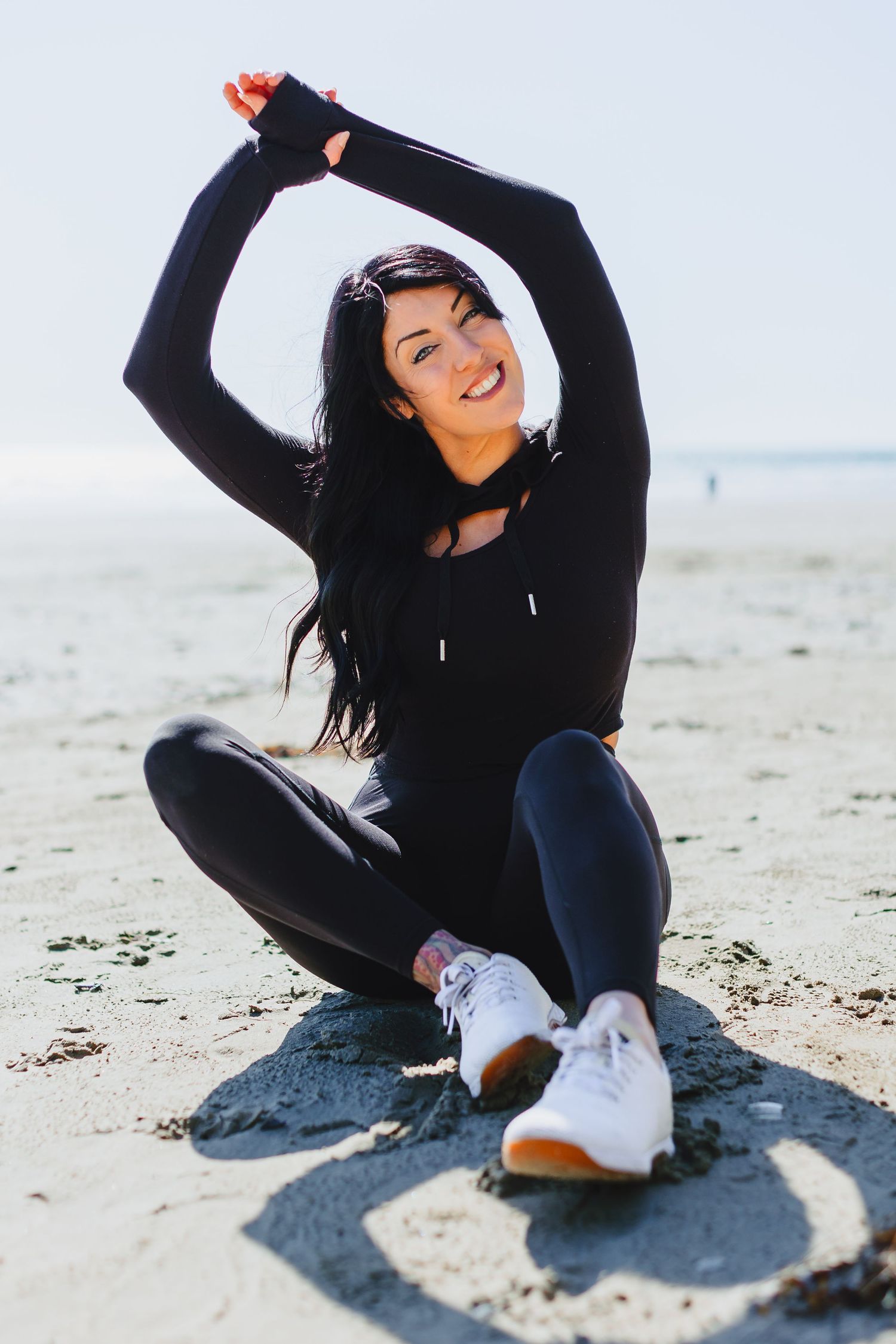 Athletic person in black workout clothes stretches and smiles during beachside fitness routine on sunny day.