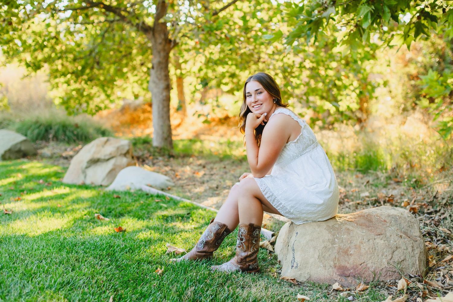 A figure in a white dress sits thoughtfully on natural stone boulders surrounded by lush grass and trees in golden sunlight.
