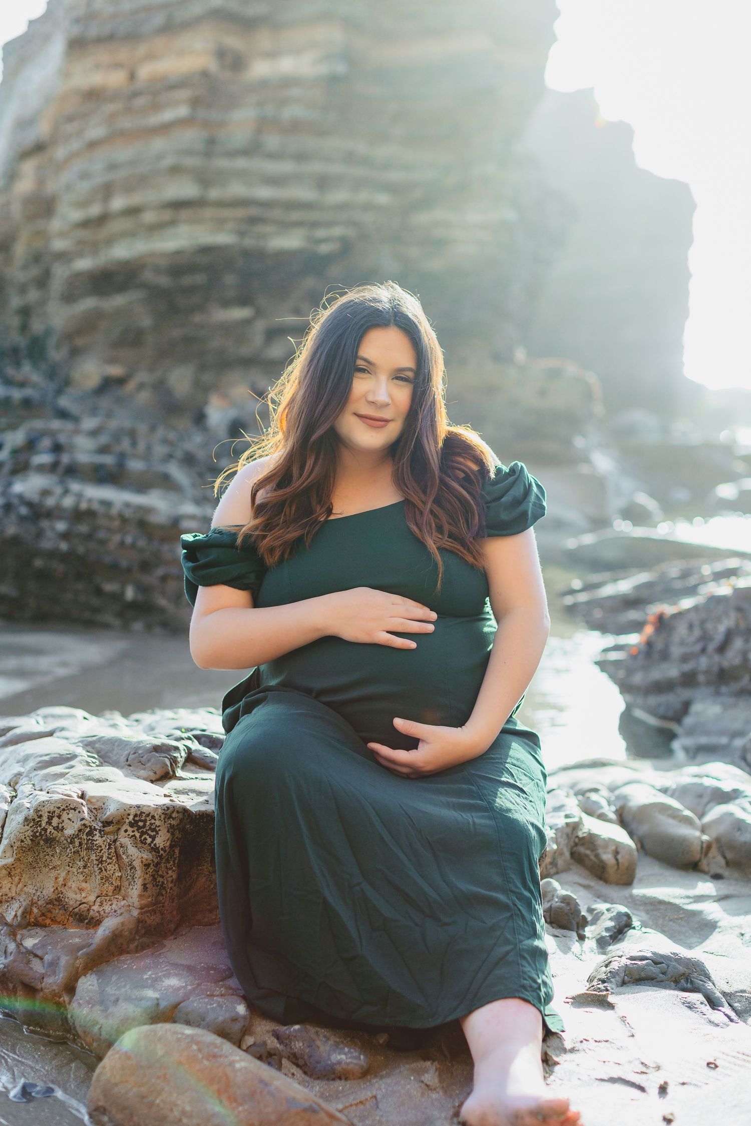 Maternity portrait at rocky beach with dramatic cliff backdrop and ocean waves.