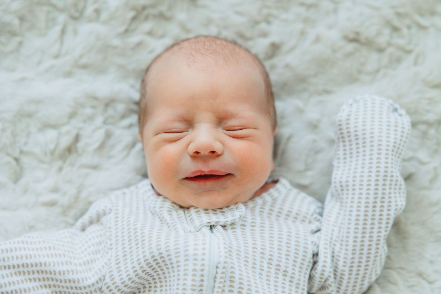 A smiling newborn baby wearing a white knit sweater lies on a cream colored background.