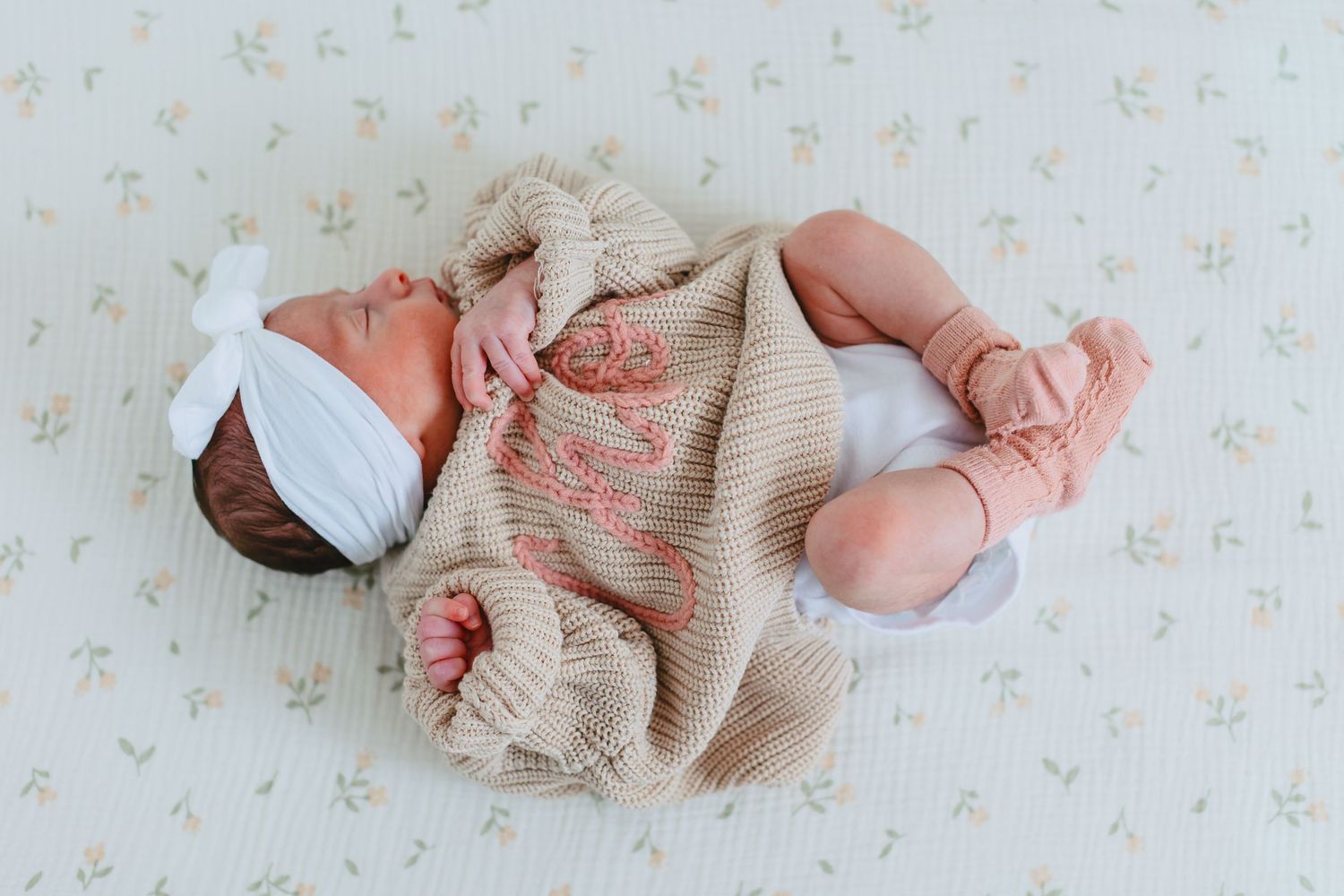 Newborn baby stretching while wearing a beige knit outfit on floral background.