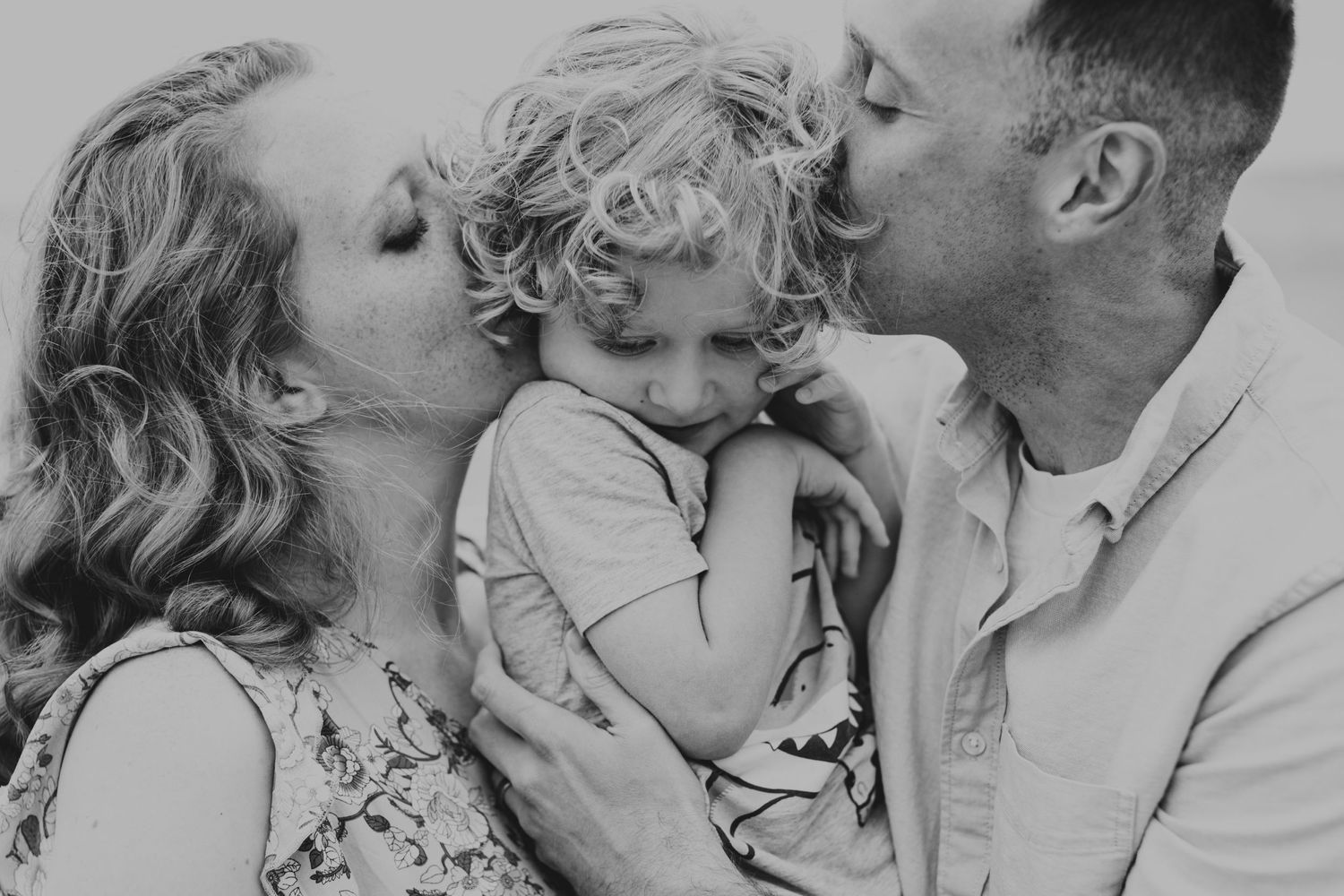 Black and white portrait of parents lovingly kissing their young child from both sides in an intimate family pose.
