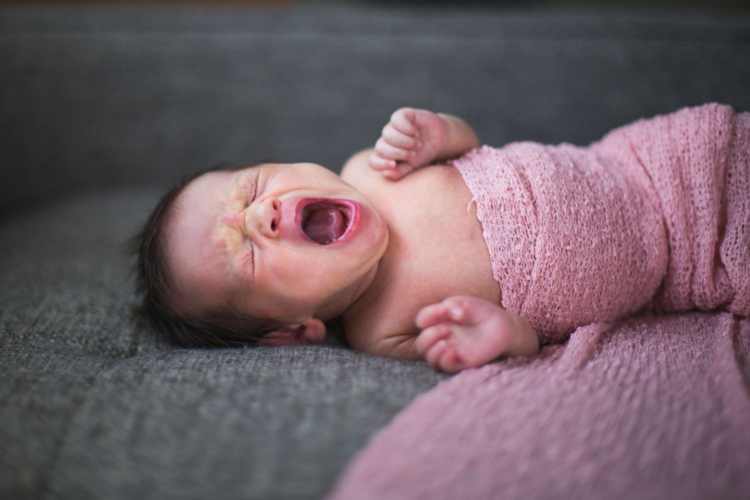 Baby wrapped in pink knit blanket crying on gray couch.