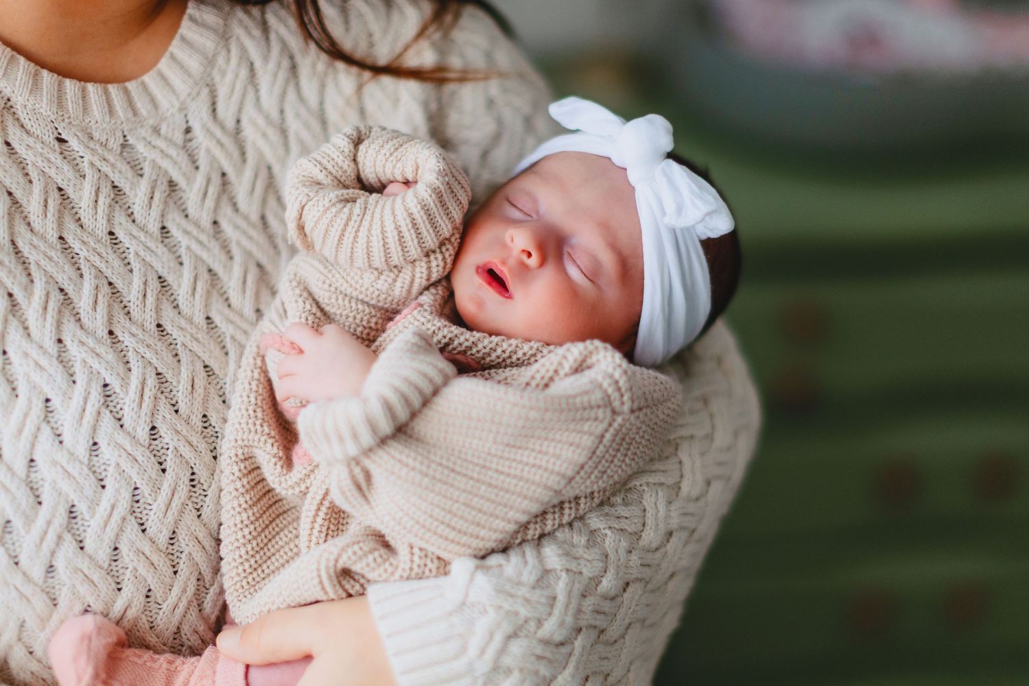 Sleeping newborn in beige knitted outfit and white bow being cradled.