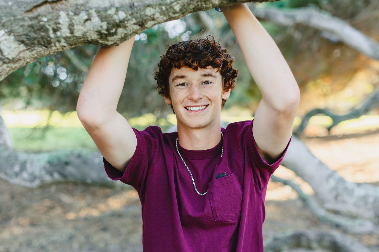 A person in purple shirt poses playfully under a tree branch outdoors.