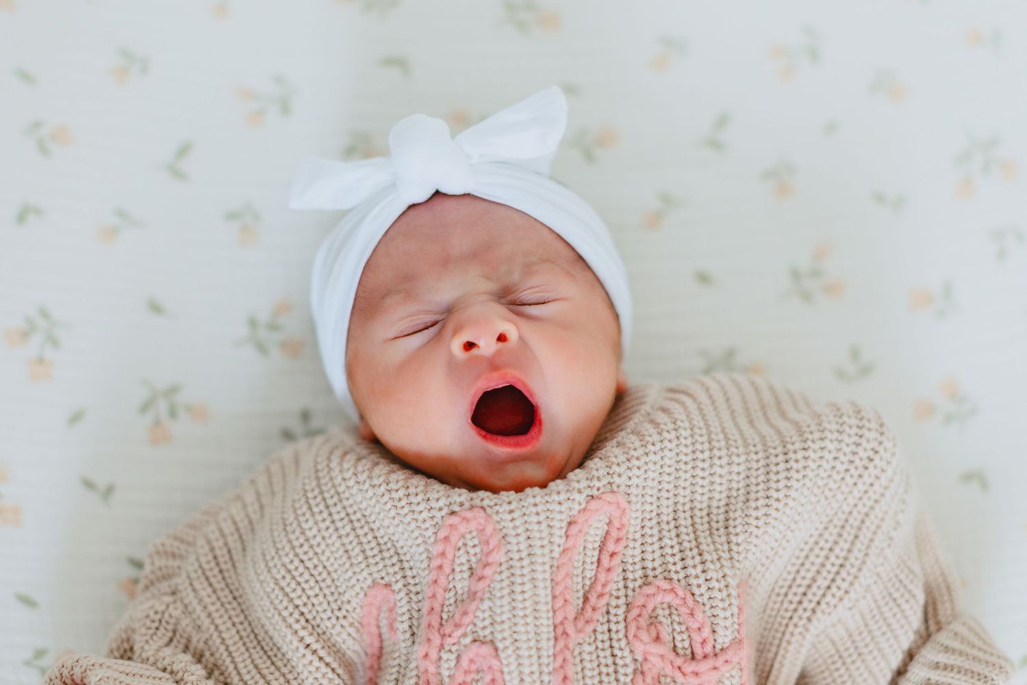 Newborn baby yawning while wearing a white bow headband and beige knit sweater.