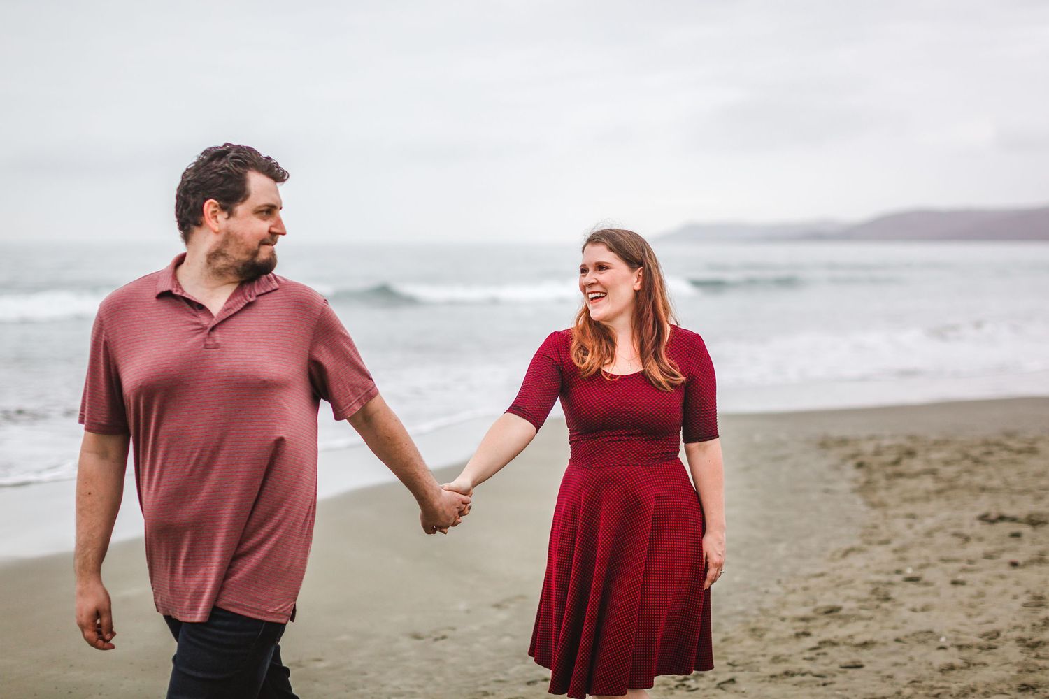 A couple walks hand in hand along a misty beach wearing casual attire.