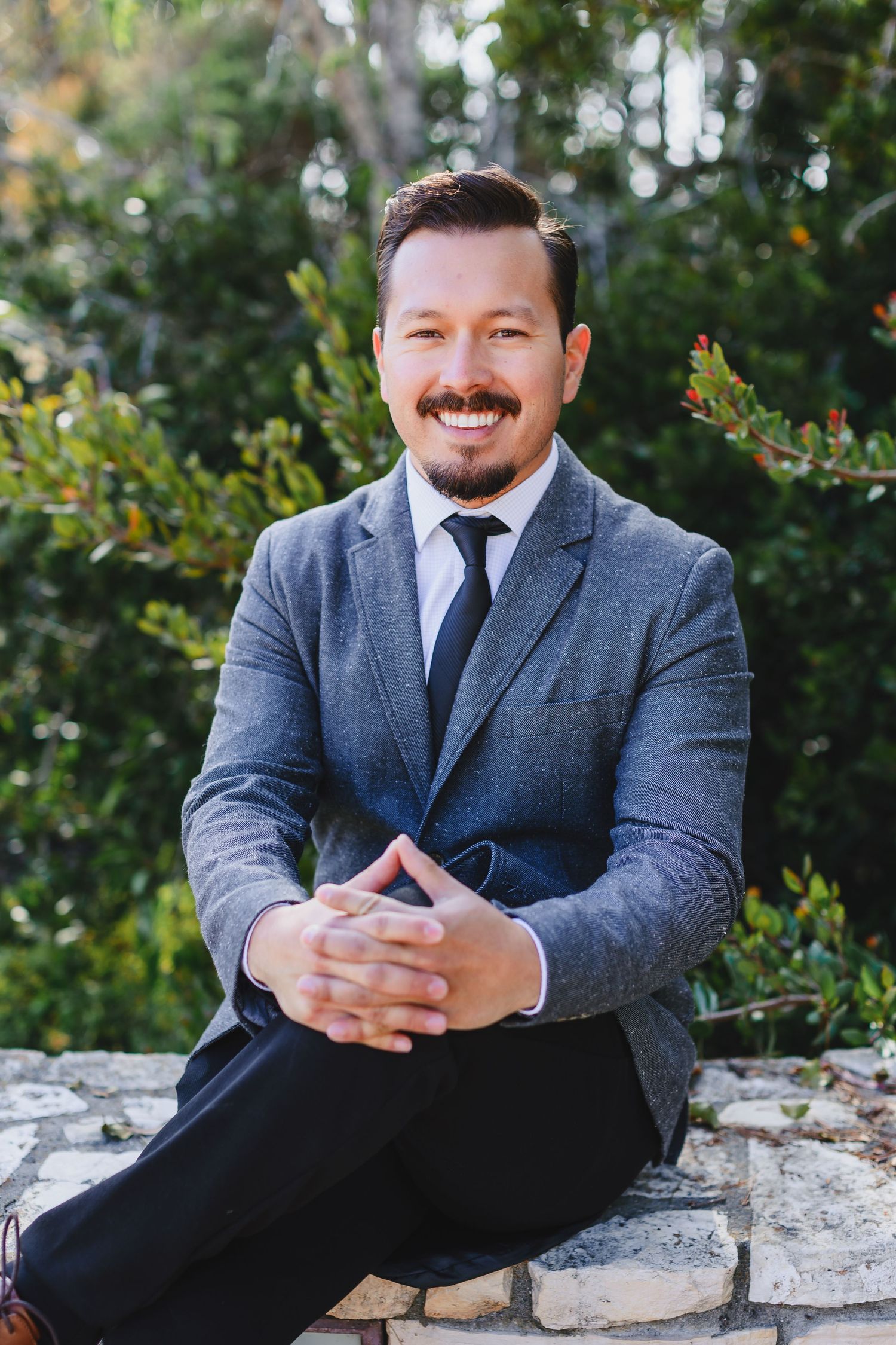 Professional headshot of person in gray suit sitting on stone wall with greenery behind.