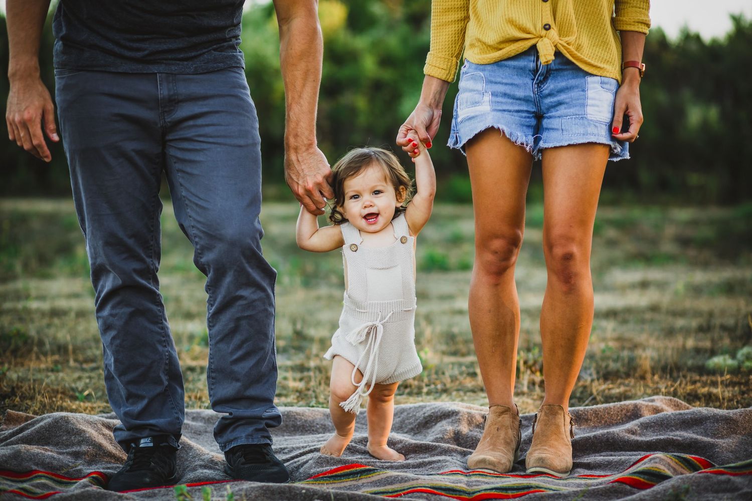 A joyful toddler walking on a blanket while holding hands with parents outdoors.