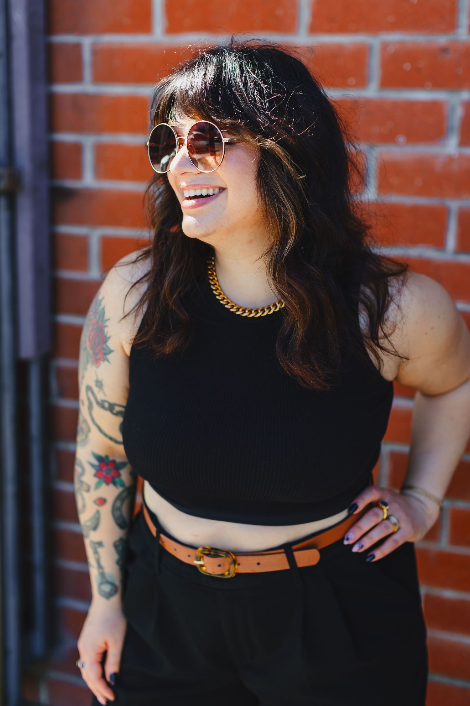 Stylish woman in black crop top with tattoos poses against brick wall.
