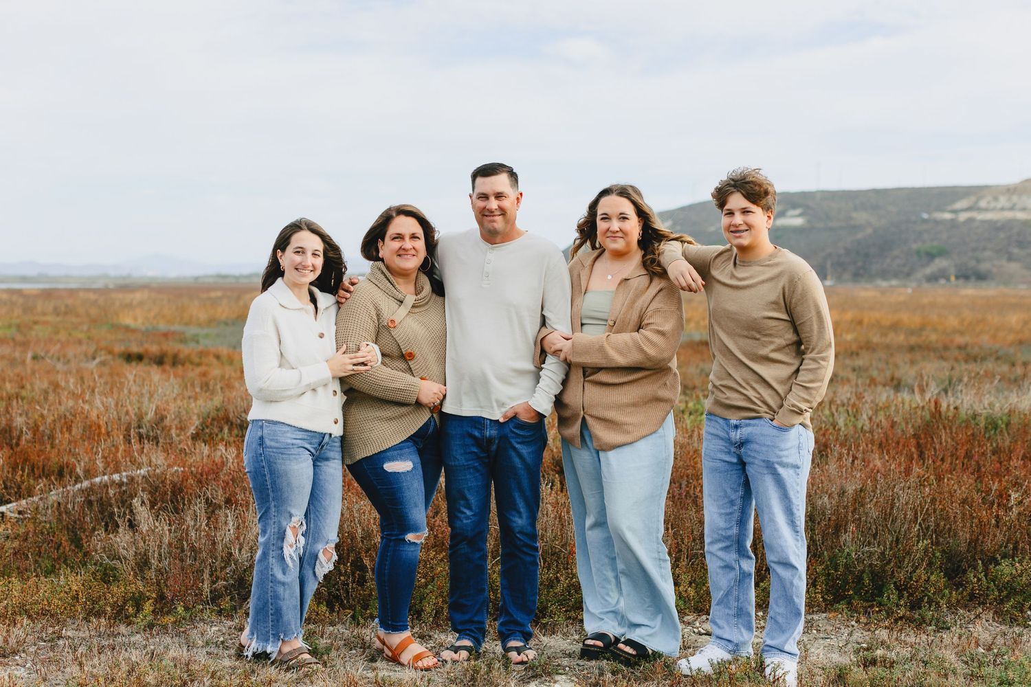 Family group in coordinated neutral outfits pose together against a rustic outdoor backdrop.