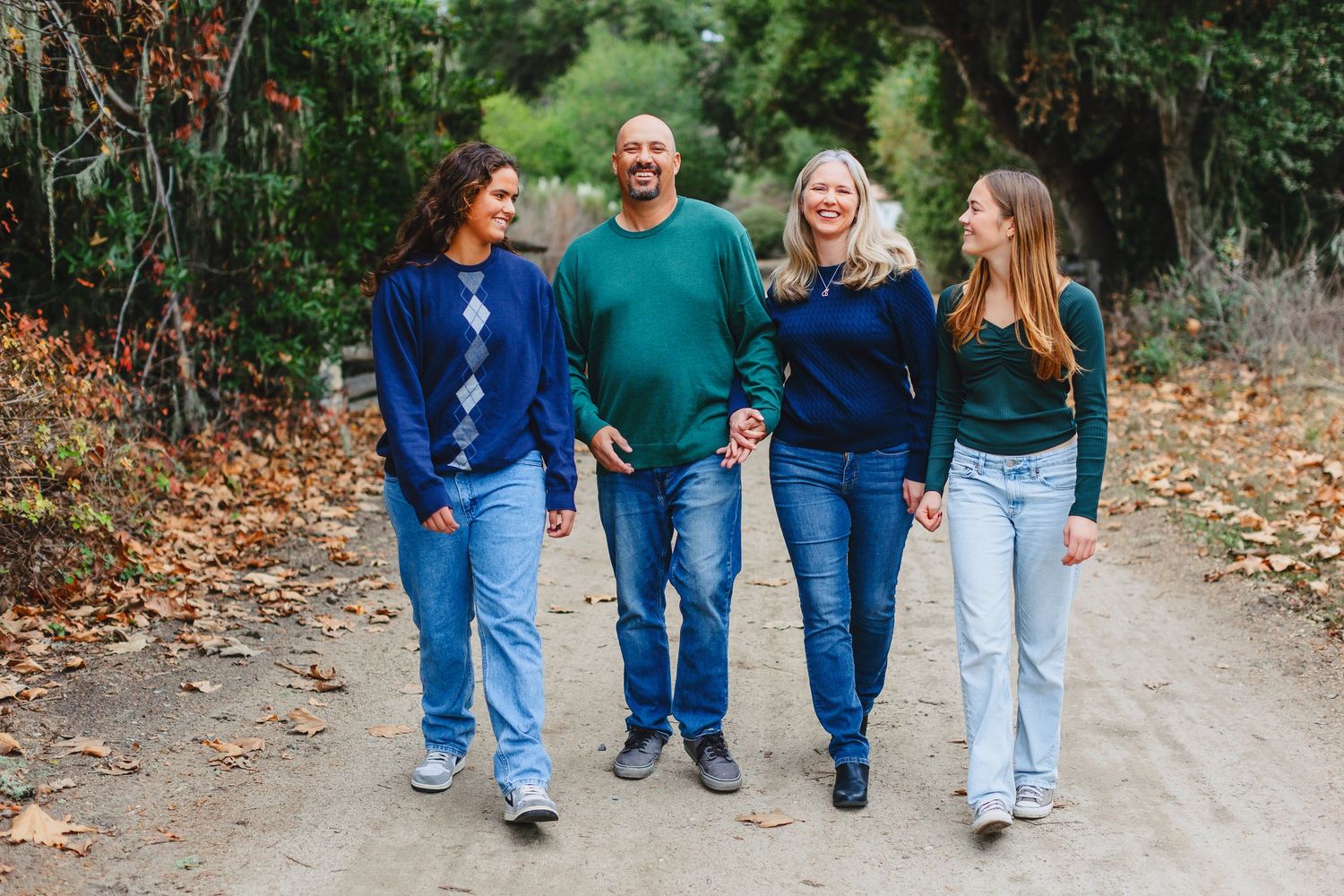 Four people walking together on a dirt trail wearing coordinated blue and green sweaters and jeans in autumn.