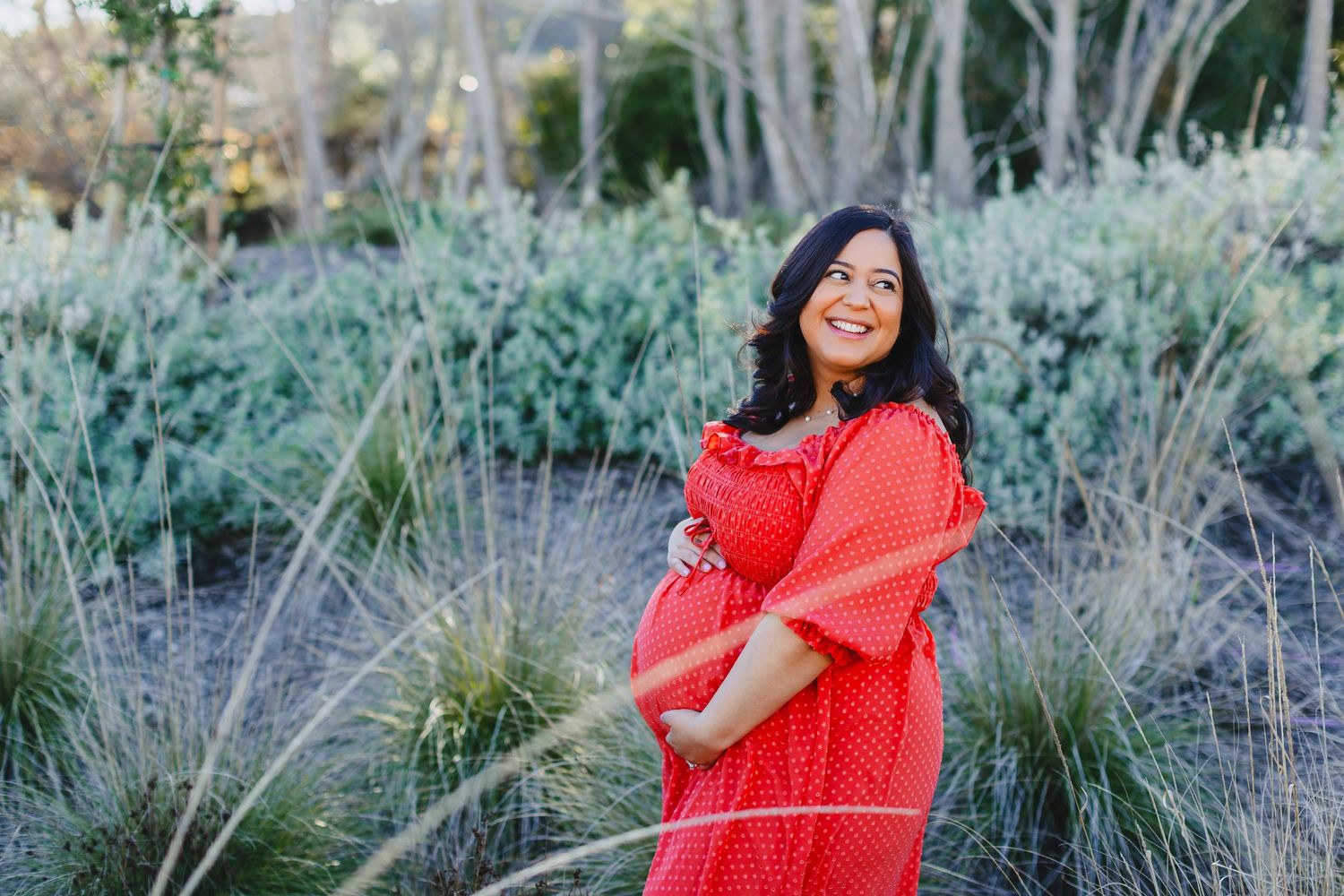 Expectant mother in flowing red dress poses in garden setting with ornamental grasses.