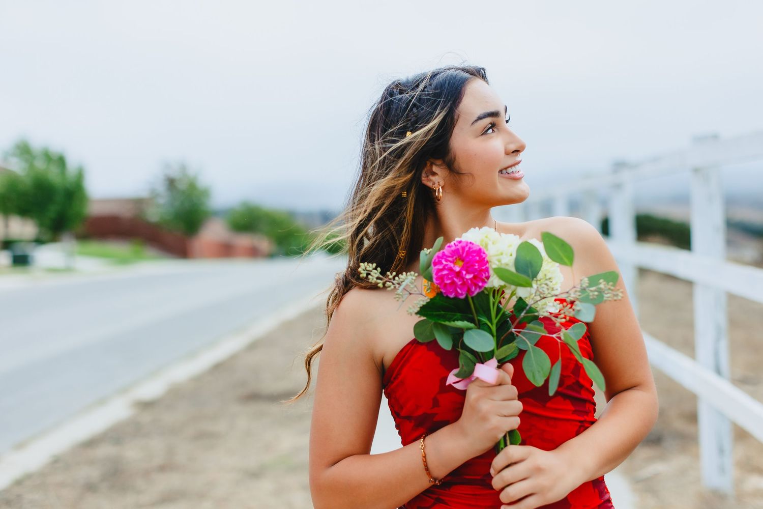 A person in a red dress holds a bouquet of pink roses while looking off into the distance.
