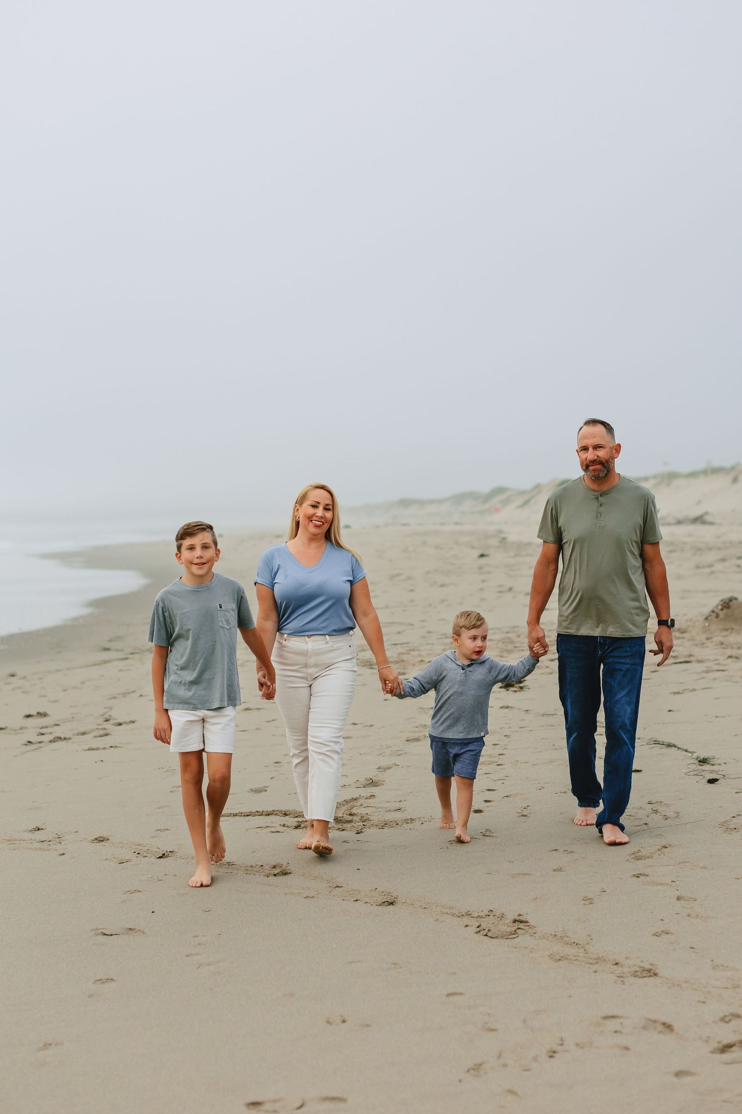 Family of four walking hand in hand along a misty beach shoreline.