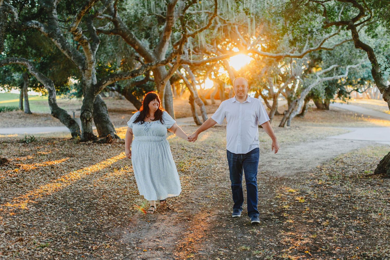 Couple walking hand in hand through sun-dappled grove at sunset.