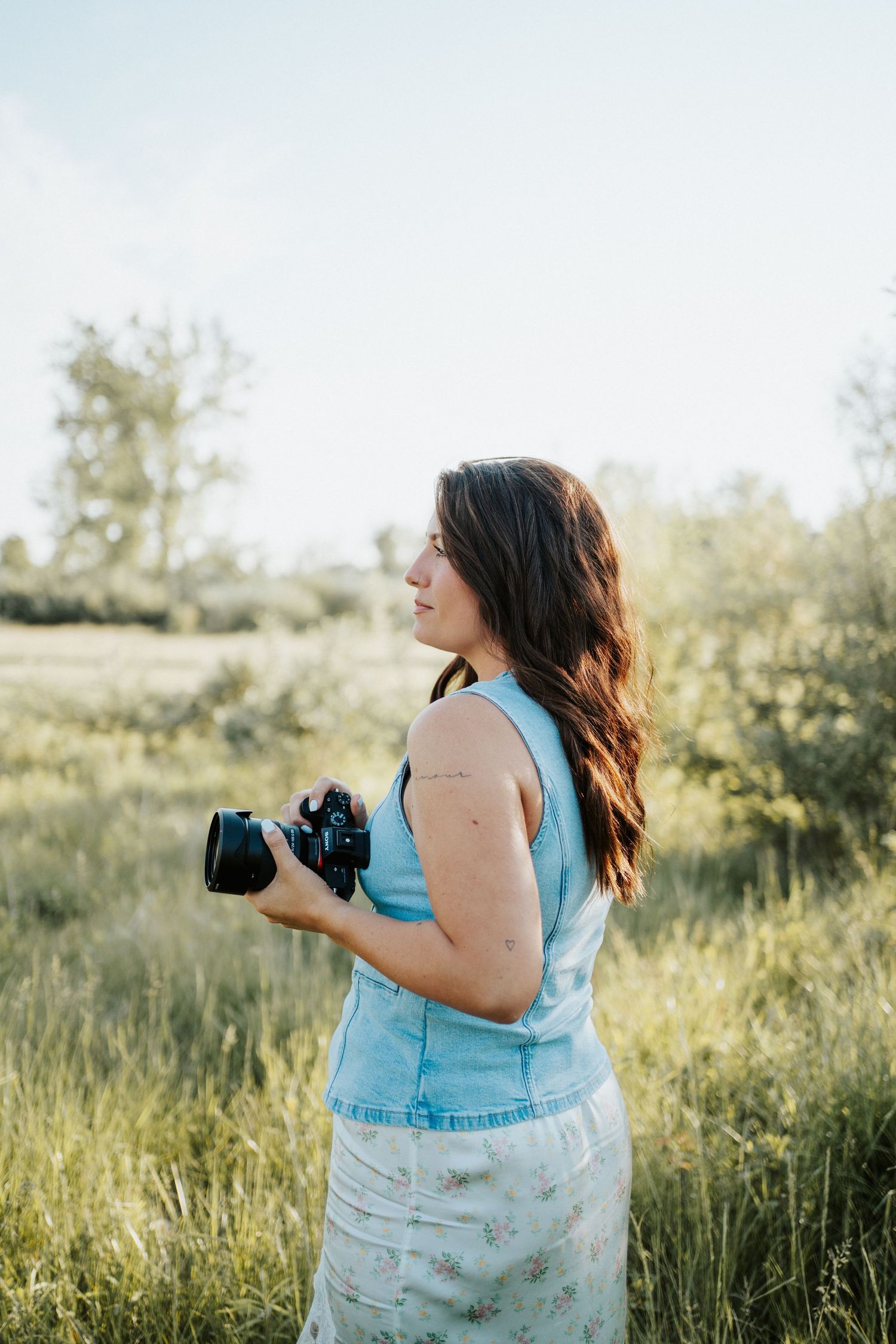 Photographer in blue denim vest and floral skirt holds camera in sunlit meadow during golden hour.
