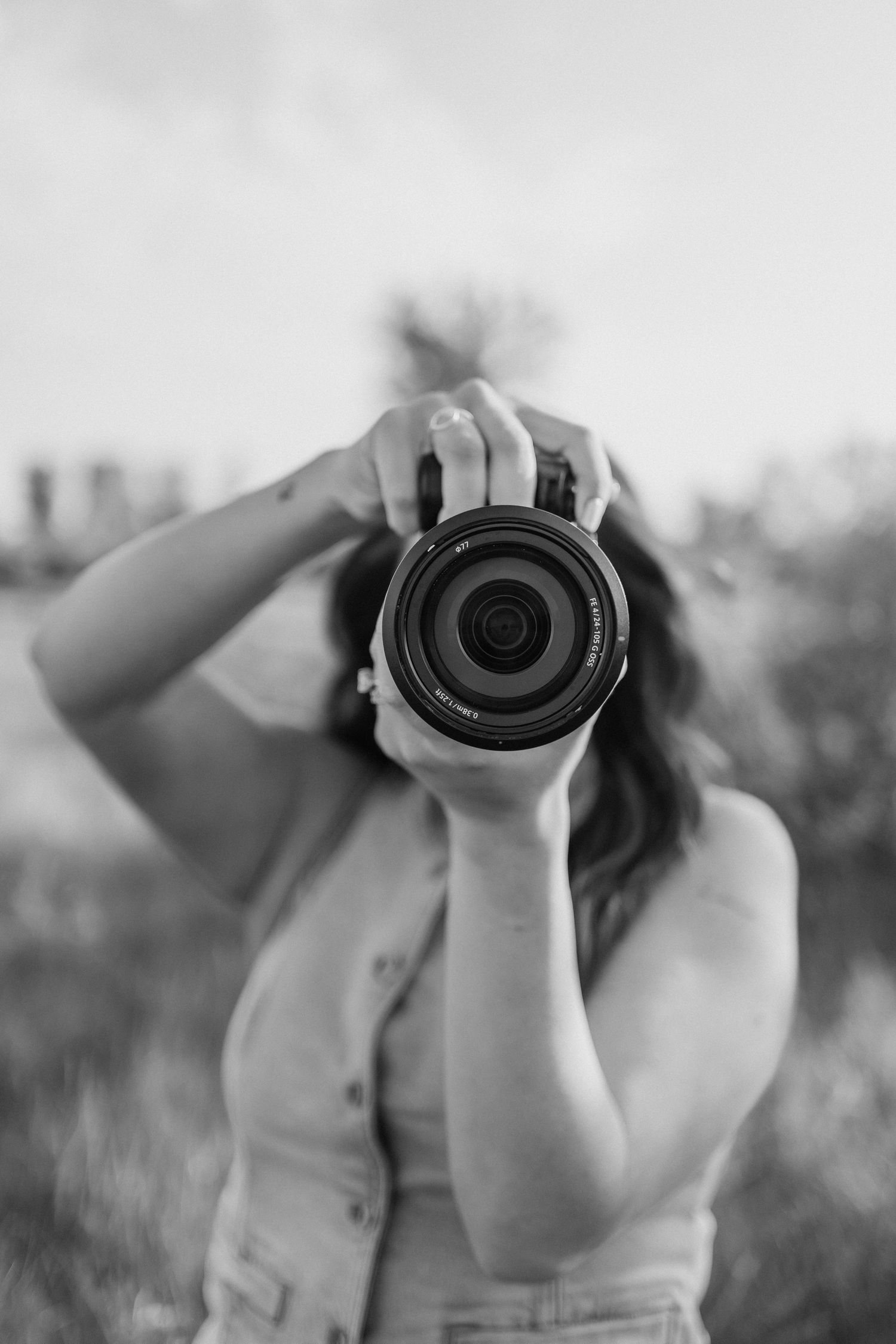 Black and white photo of photographer holding a digital camera with large lens against blurred outdoor background.