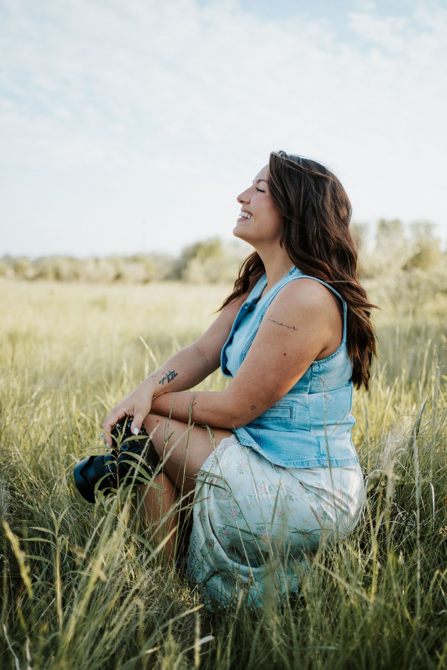 A photographer sits in tall grass wearing a blue tank top and denim shorts while laughing in golden sunlight.