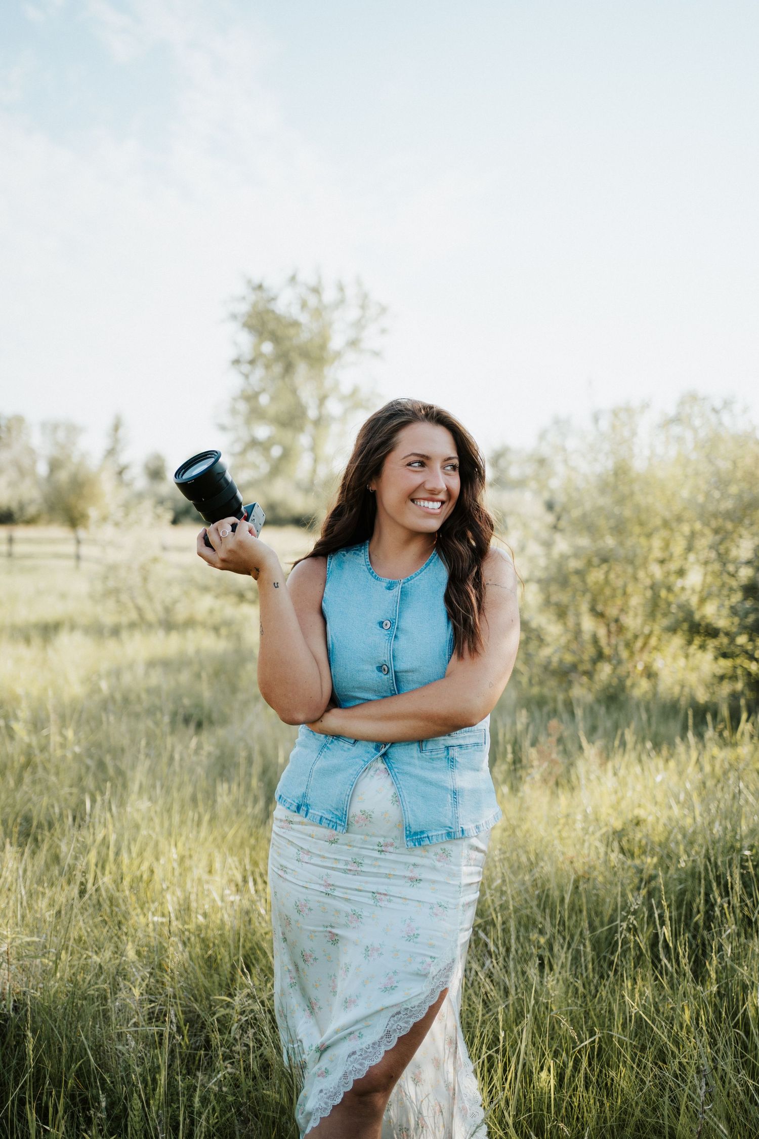 A photographer in a blue sleeveless top and white skirt stands smiling in a sunlit meadow holding a camera.