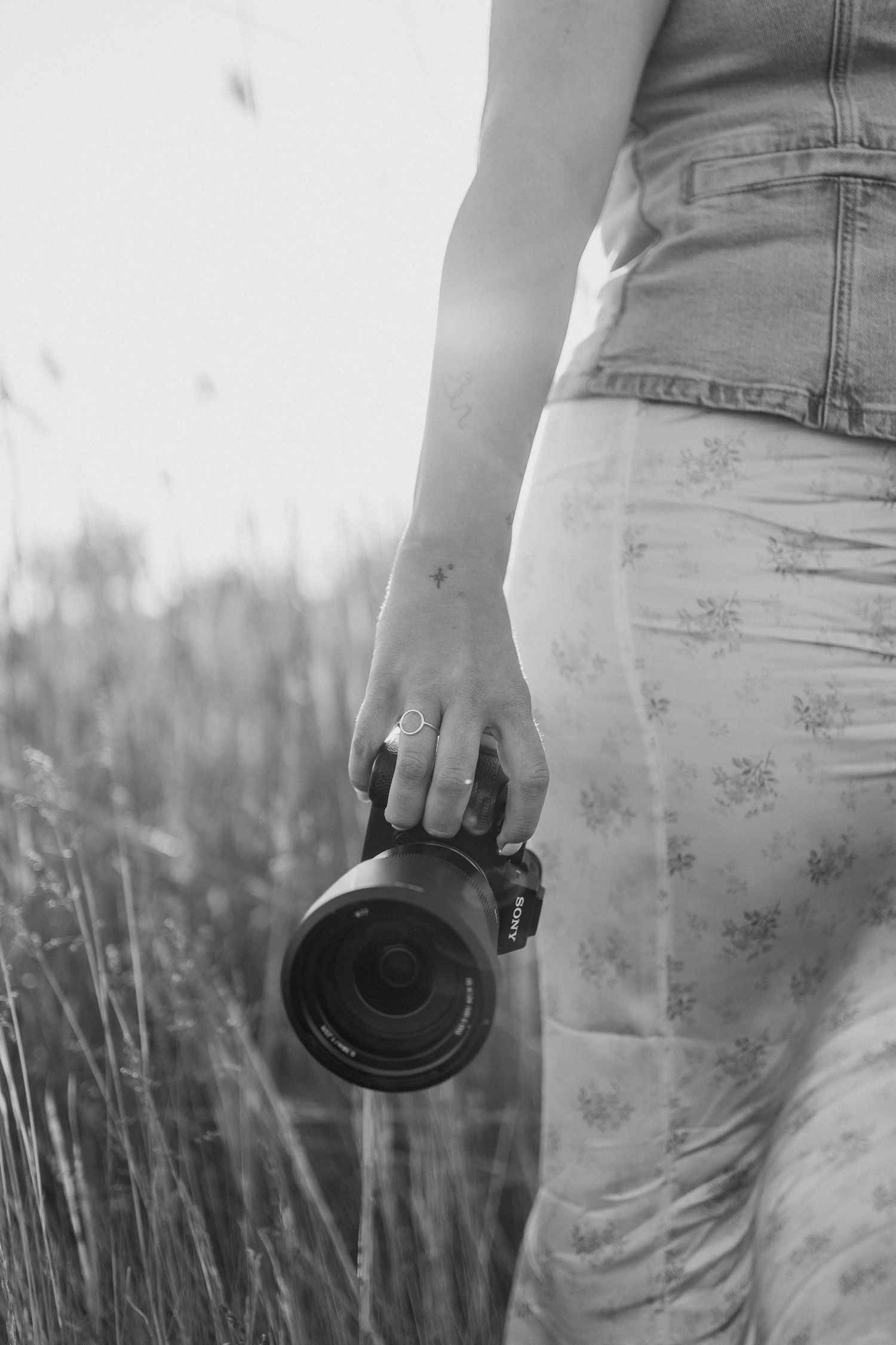 Black and white photograph of someone holding a camera lens while standing in a field of tall grass at sunset.
