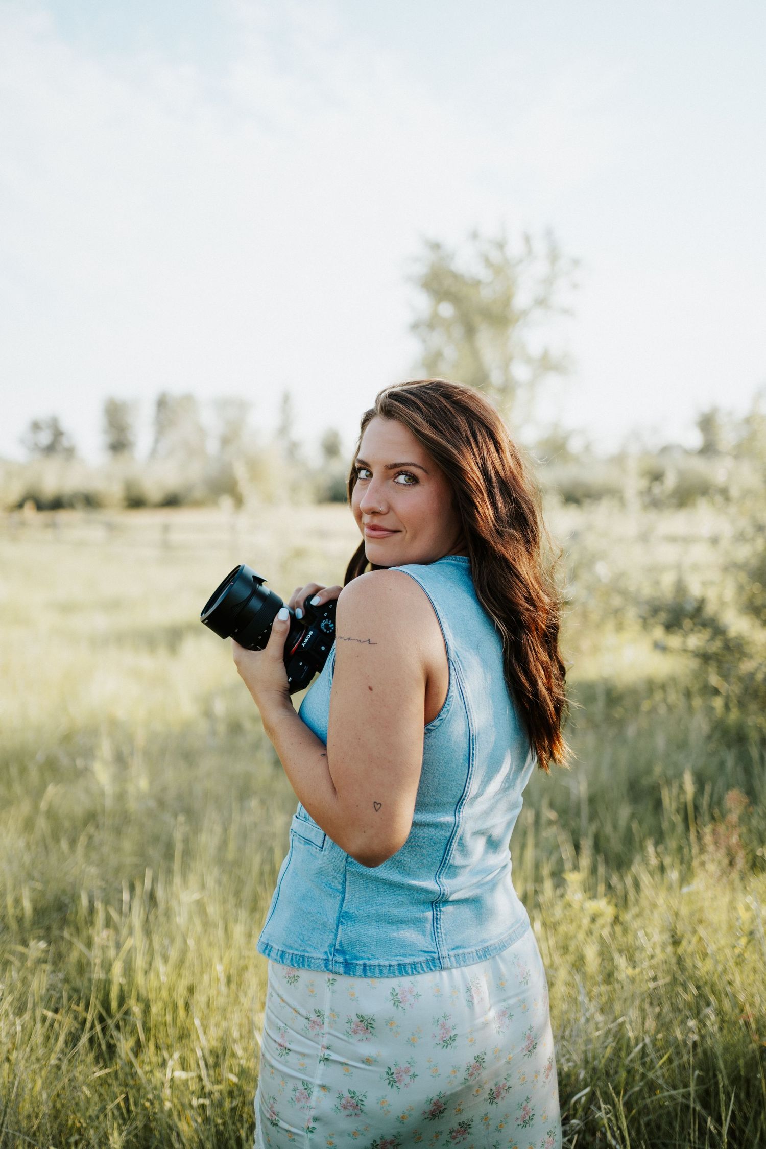 A photographer in a light blue top and floral skirt holds a camera while standing in a sunlit meadow.