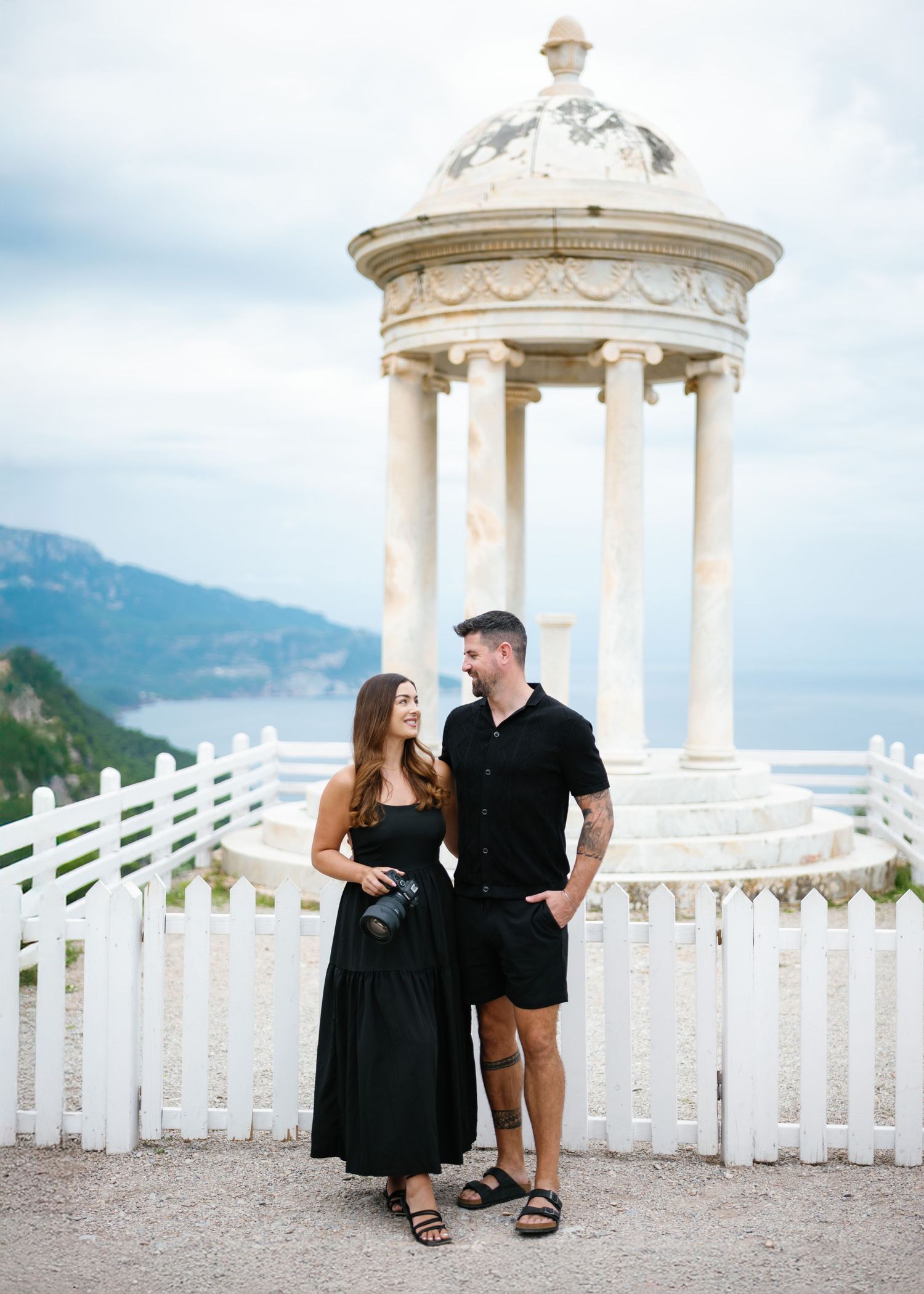 A couple in black attire shares romantic moments along a scenic coastal viewpoint with white pavilions and mountains.