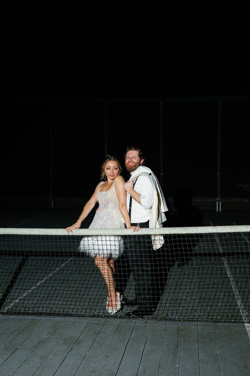 A couple poses playfully on a tennis court at night, with the bride wearing a short white feathered dress.