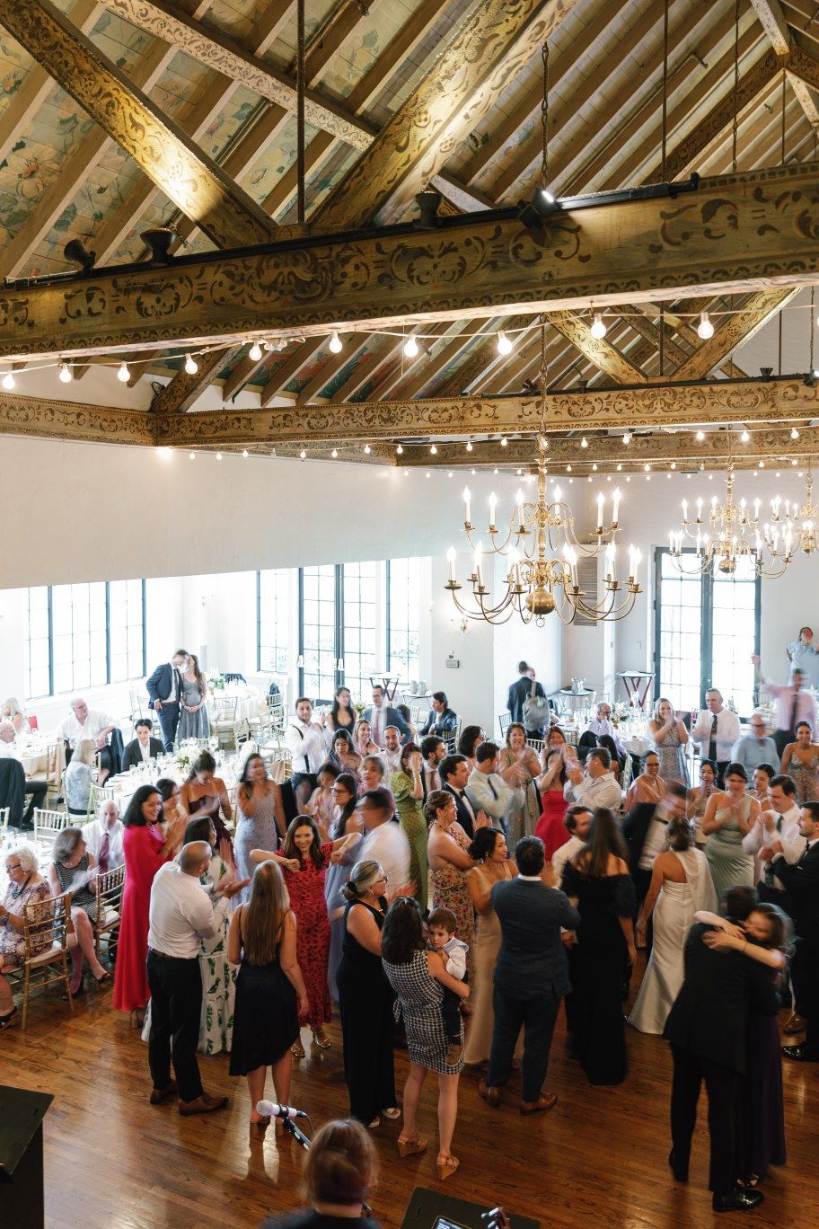 Wedding reception crowd dances in a rustic barn venue with chandeliers and exposed wooden beams.