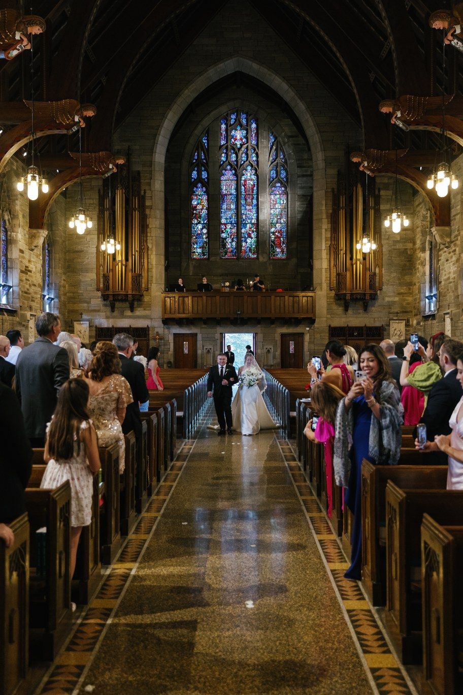 Wedding procession walks down the center aisle of a historic church with gothic architecture and stained glass.