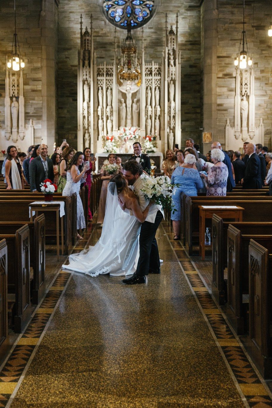 A couple stands in the center aisle of a grand church during a wedding ceremony as guests watch from wooden pews.