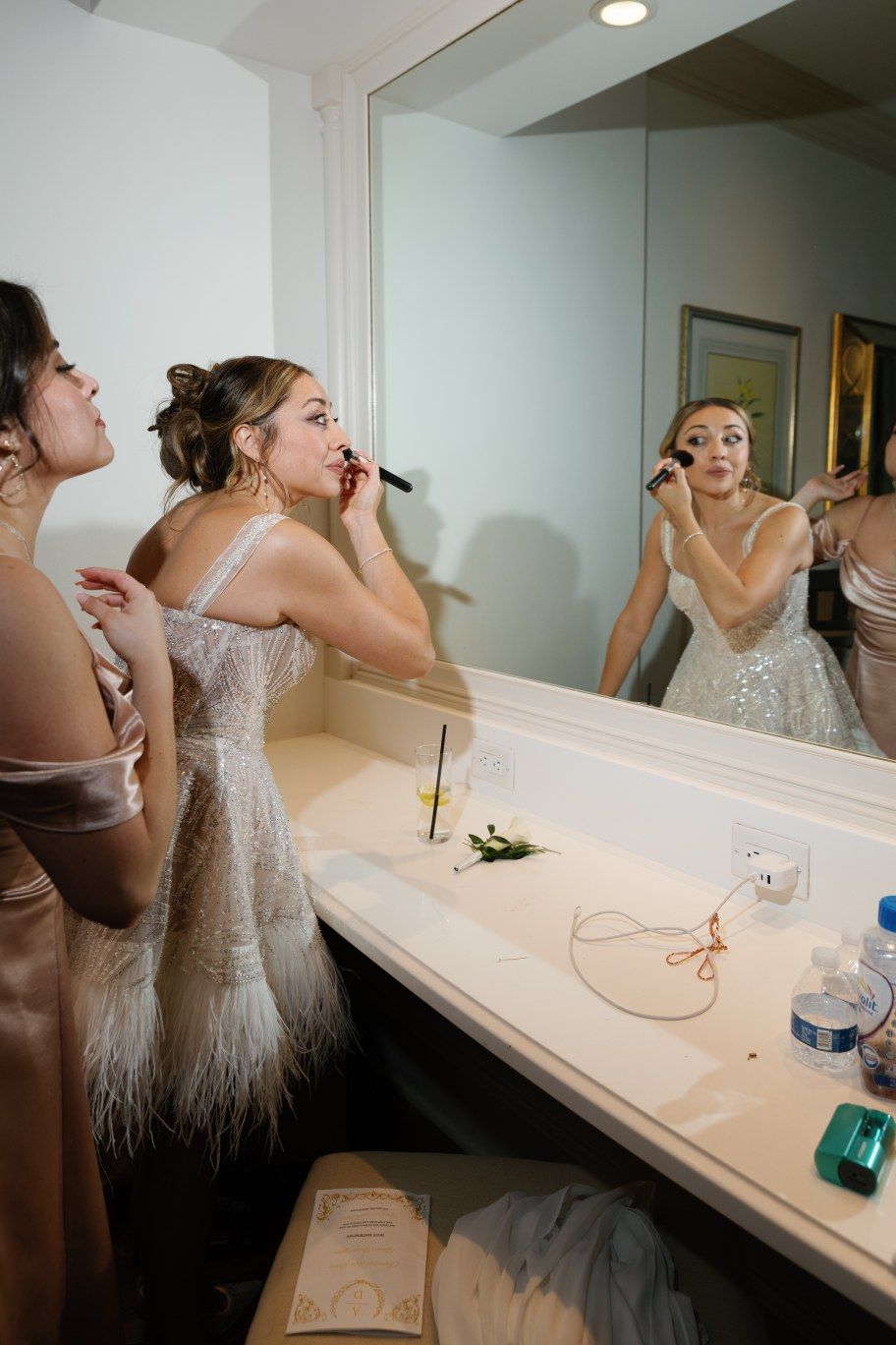 Two people in elegant dresses apply makeup at a vanity mirror counter during formal event preparations.