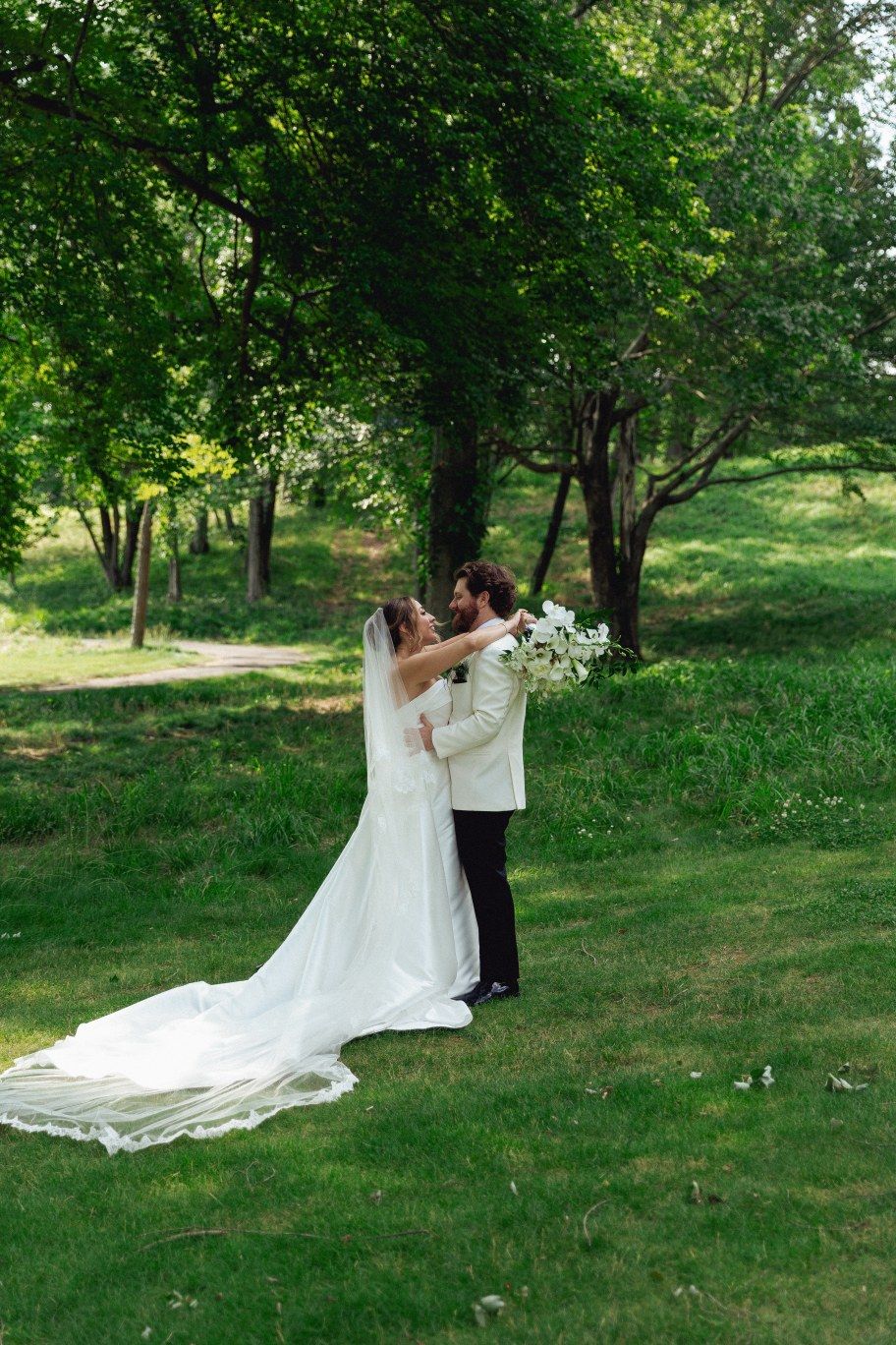 A couple in wedding attire dances on a lush green lawn surrounded by trees on their special day.