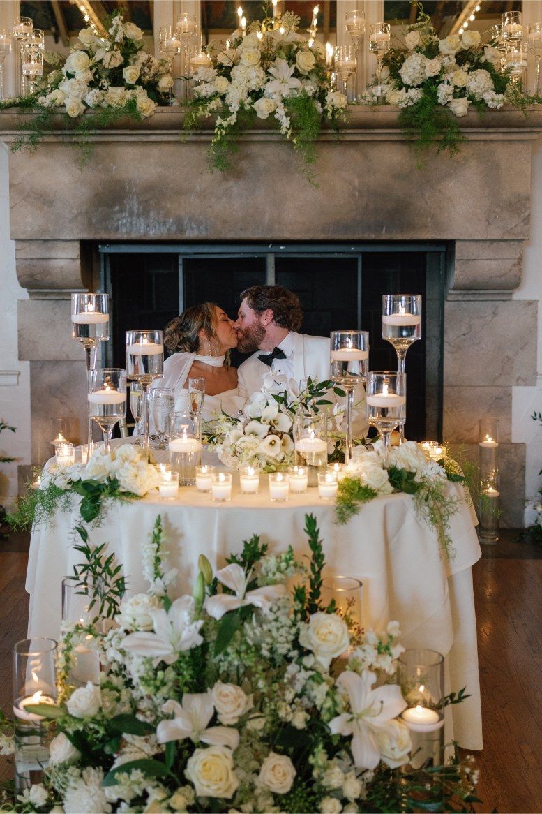 Romantic sweetheart table decorated with white flowers and candles in front of a stone fireplace.