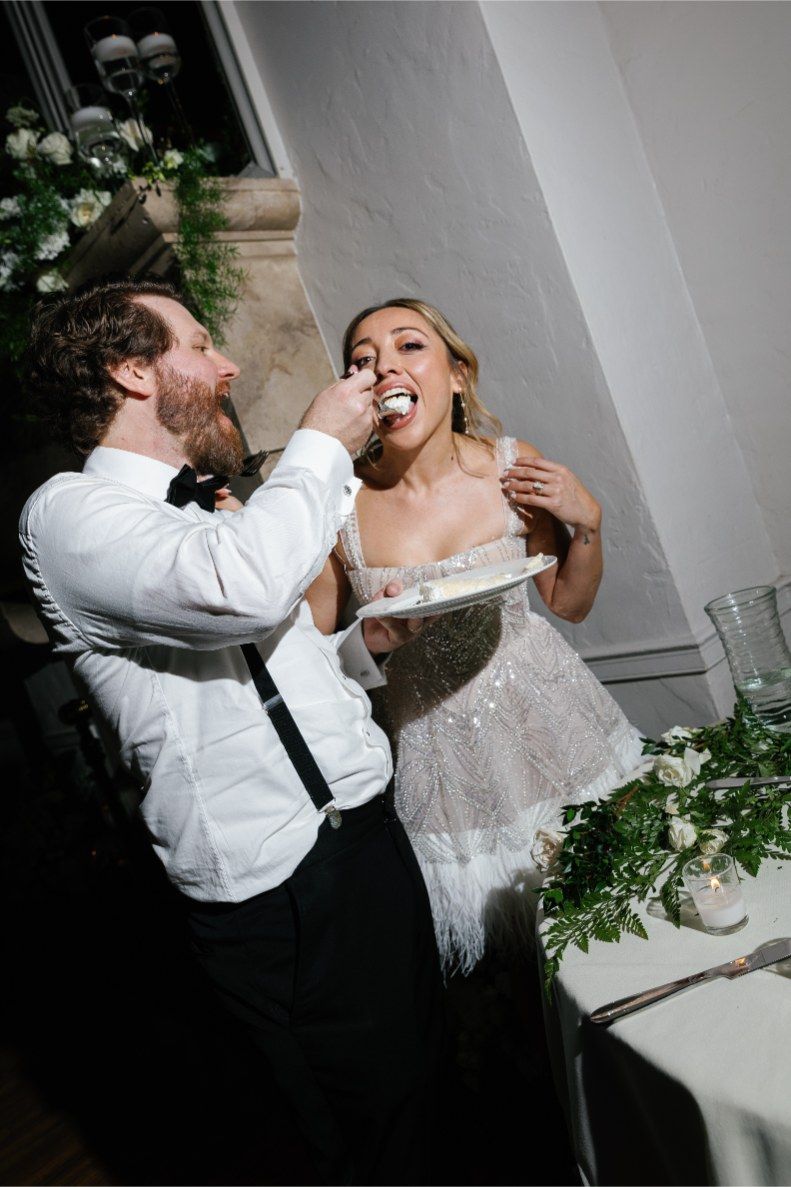 A couple playfully feeds each other wedding cake at their reception with white floral decorations in the background.