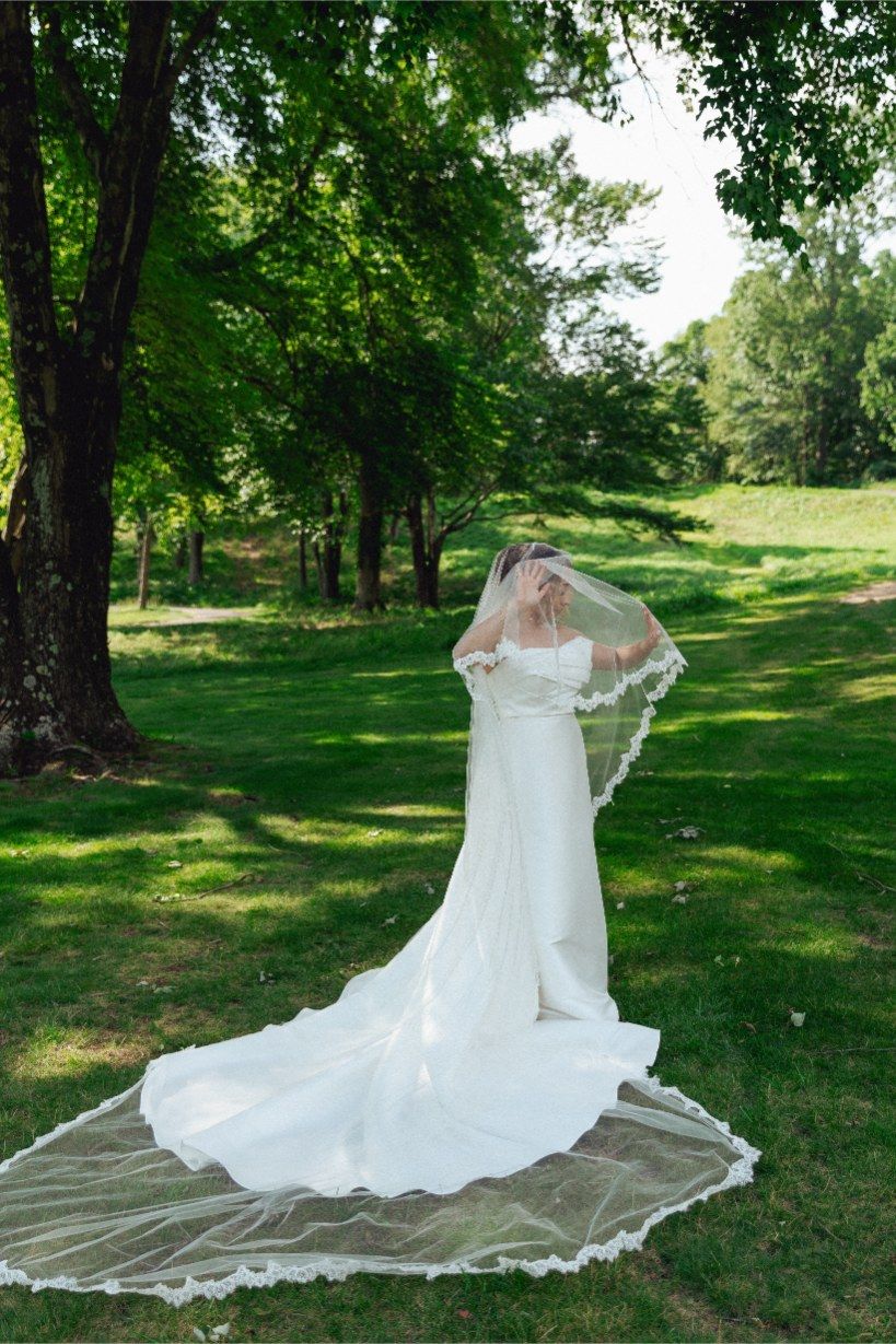 A radiant figure in a flowing white wedding gown with a long veil stands on a wooden platform in a lush green garden setting.