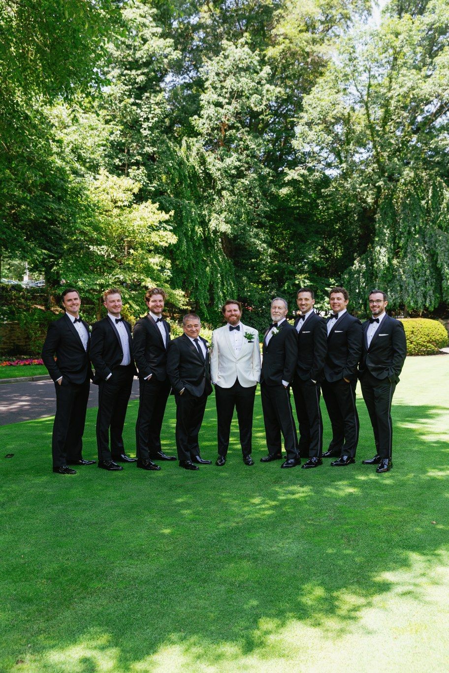 Wedding party in black suits and a single white suit pose together on a manicured lawn with green trees in background.