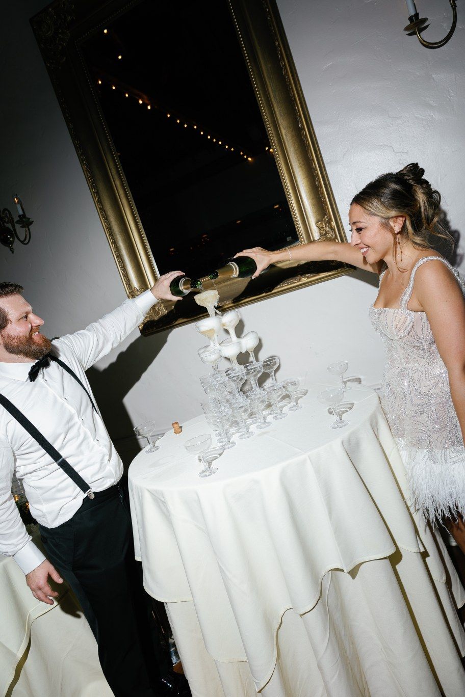 A playful moment at a wedding reception as two people reach across a decorated table with champagne glasses.