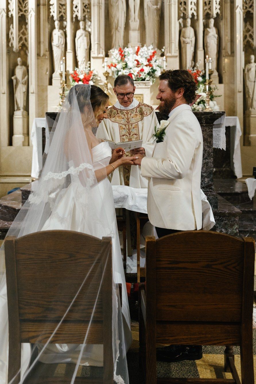 A traditional church wedding ceremony takes place at an ornate altar adorned with red and white floral arrangements.