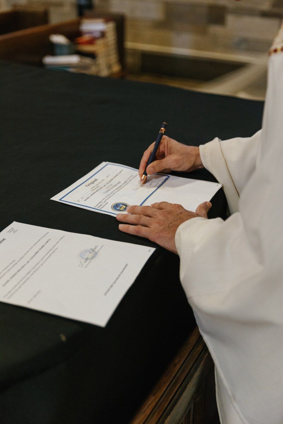 Person in white shirt signing legal documents on a dark table surface while holding a pen.