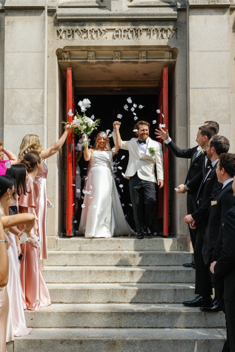 Newlyweds celebrate with guests throwing flower petals as they exit a church doorway with red pillars down stone steps.