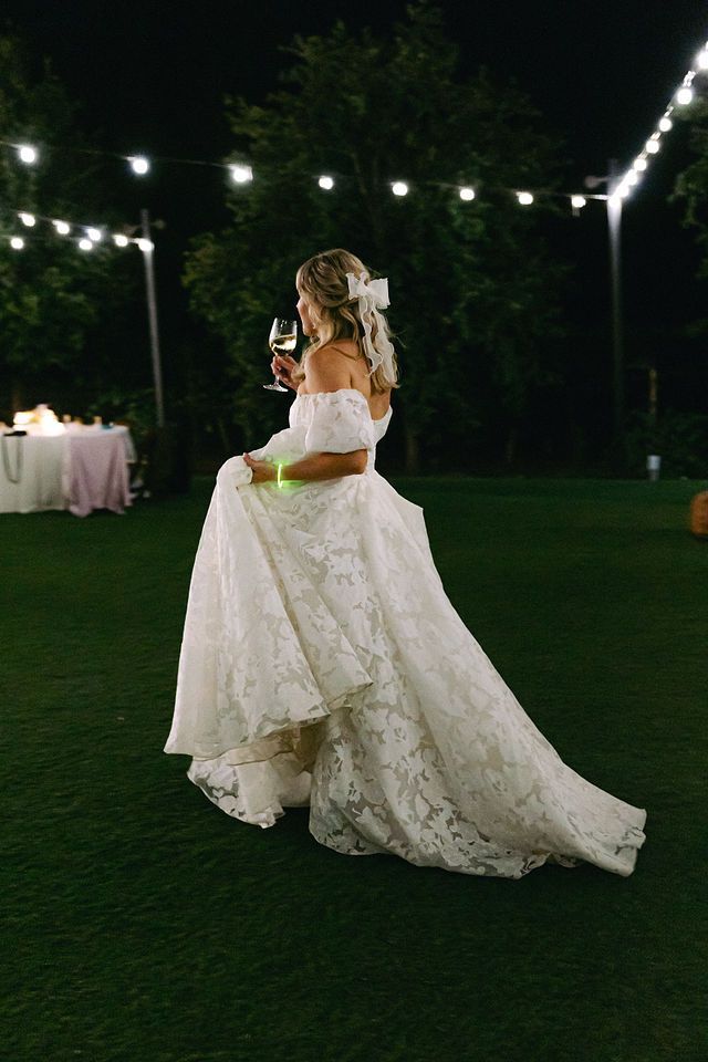 Bride in lace gown holds wine glass on illuminated lawn at night.