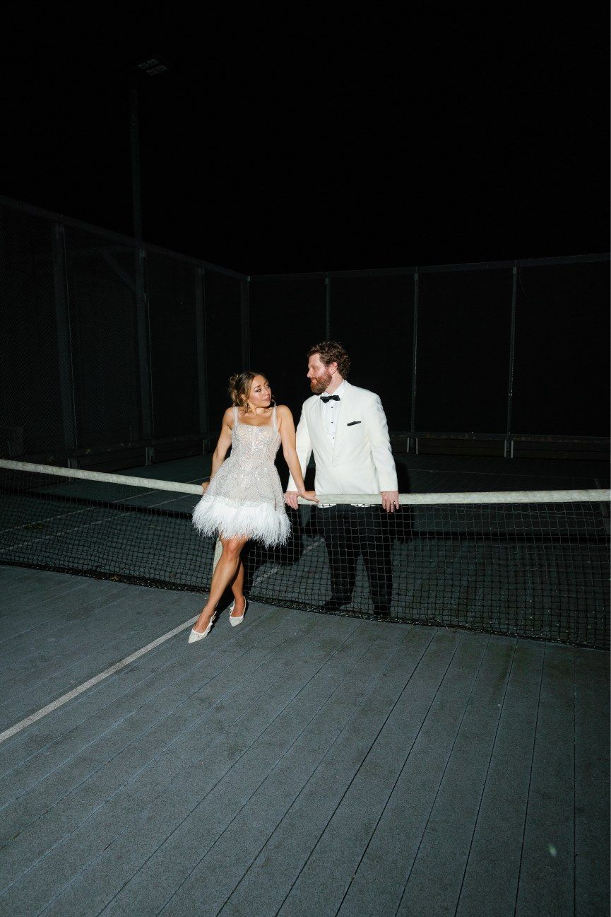 A couple in wedding attire stands together on a dark tennis court at night, the bride wearing a short feathered dress.