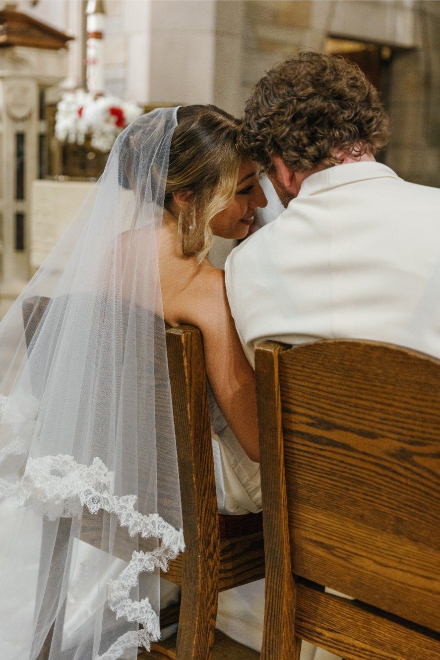Bride wearing a long white veil leans against wooden church pews during a wedding ceremony.
