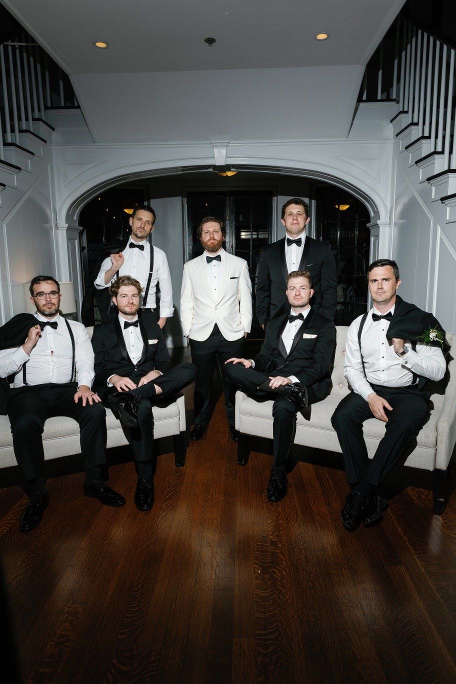 Wedding groomsmen in black tuxedos and white bow ties pose casually seated on floor along white staircase banister.