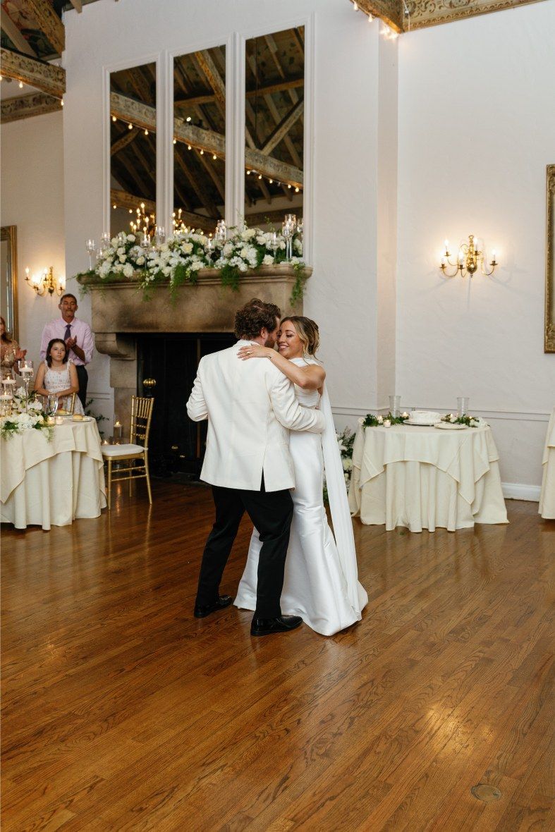 Couples dancing in an elegant white reception hall with hardwood floors and string lights.