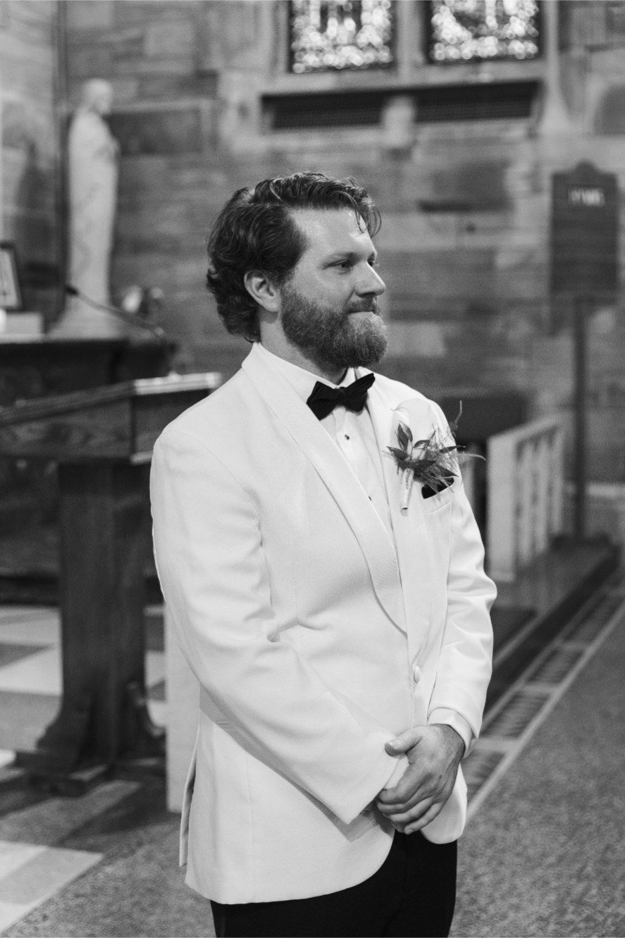 A groom in a white tuxedo jacket and black bow tie stands contemplatively in a church setting.