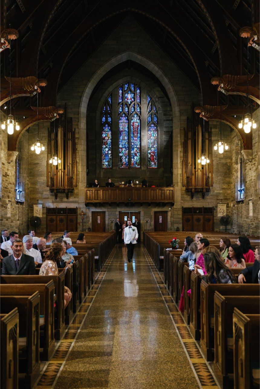Two silhouettes stand in a Gothic church aisle with stained glass windows, wooden pews and pendant lights illuminating the space.